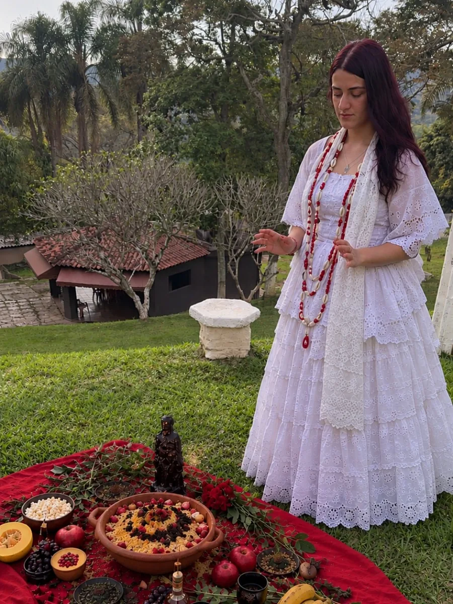 Mentora Kamily de vestido branco de renda e guias vermelhas, em pé ao lado de uma oferenda sobre pano vermelho